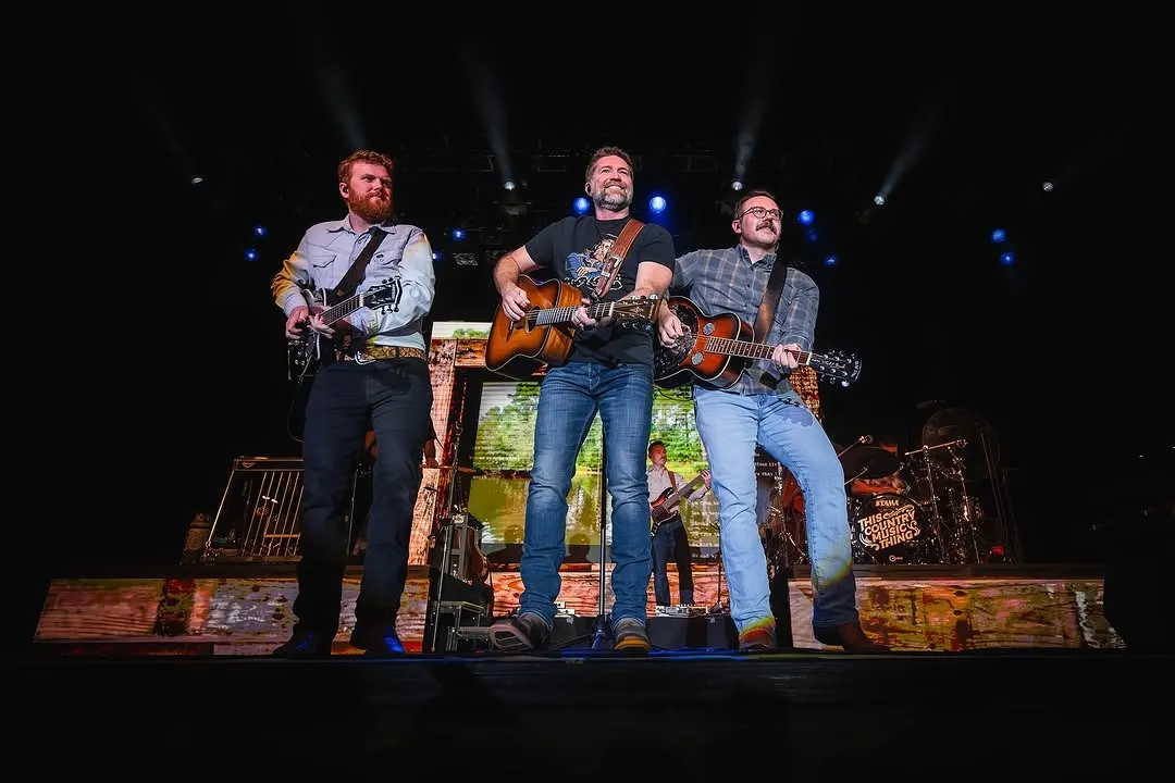 Josh Turner with two acoustic guitar players on stage in front of a pastoral / forest scene LED backdrop, 'This Country Music Thing' sign visible at side