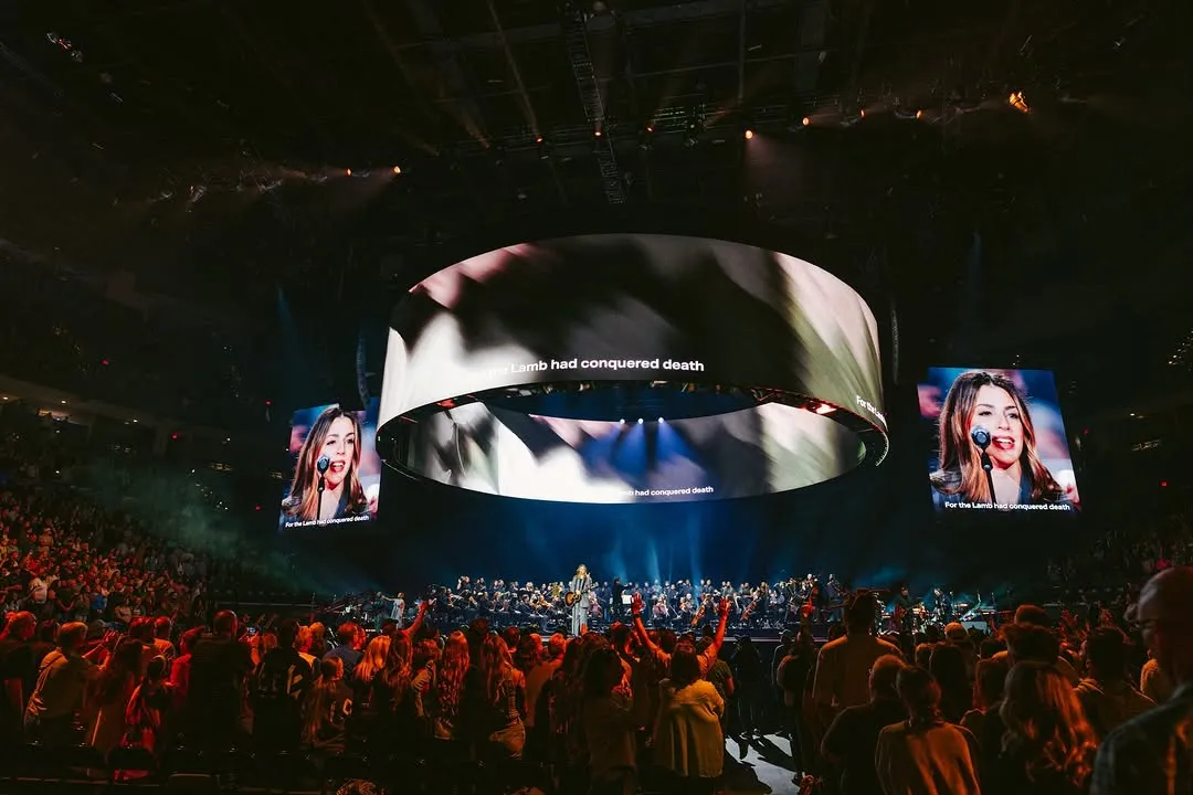 Beyond a Billion — wide arena with the 360° canopy carrying "For the Lamb had conquered death" lyrics, side IMAG screens framing a vocalist closeup, packed crowd standing