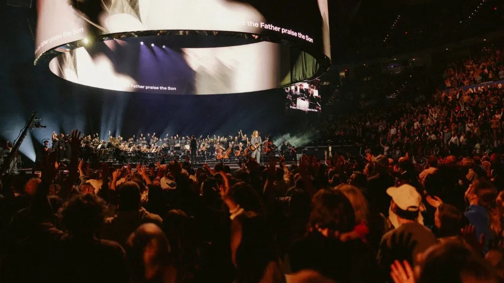 Beyond a Billion — wide arena view of the suspended 360° LED canopy with "praise the Father praise the Son" lyrics, orchestra and worship leader on stage, audience hands raised
