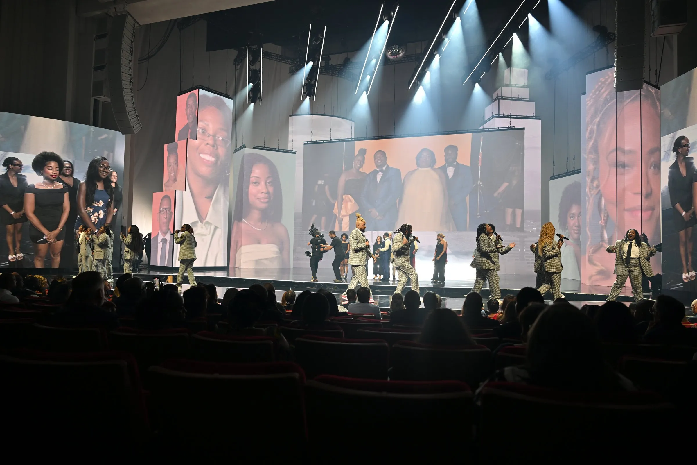 Beloved Benefit beneficiary moment — choir-like ensemble in tan suits performing on stage with portraits of program participants on the side LED panels