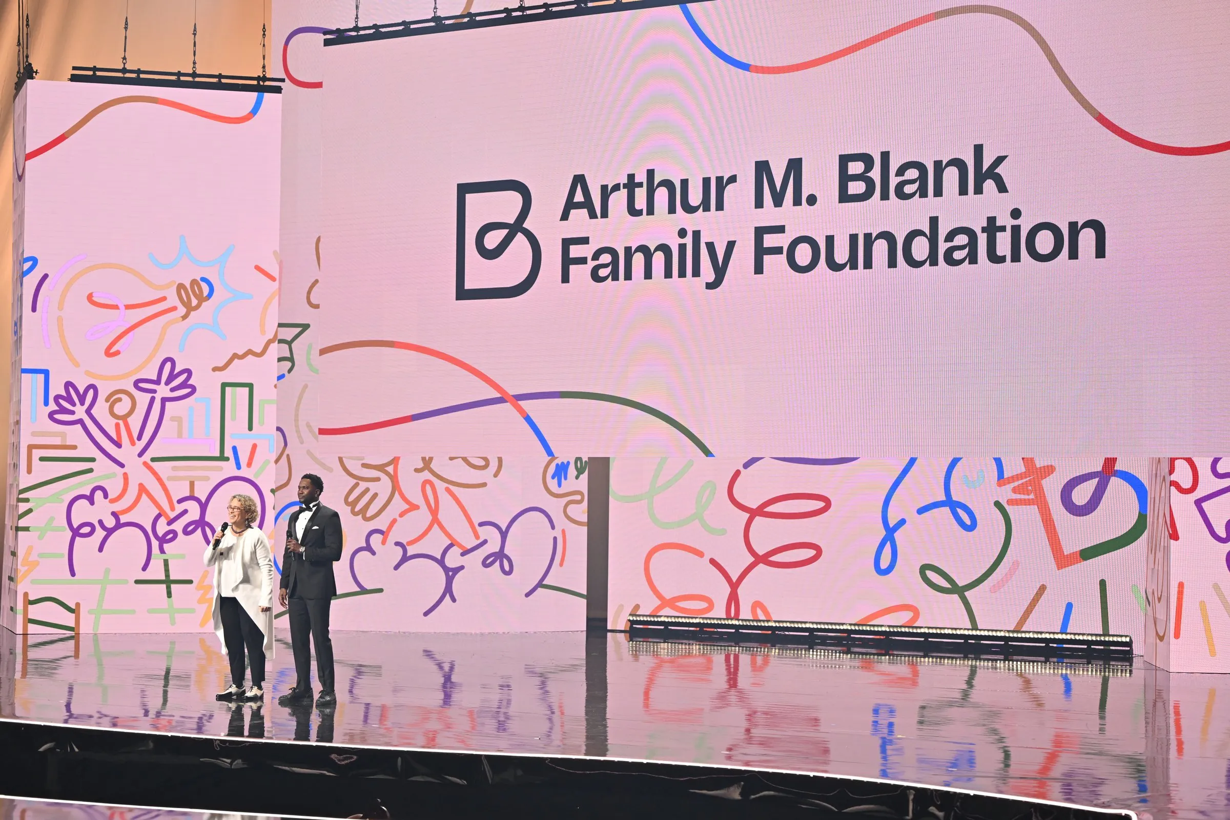 Beloved Benefit Arthur M. Blank Family Foundation sponsor moment — branded LED backdrop with abstract scribble illustration, two presenters on stage