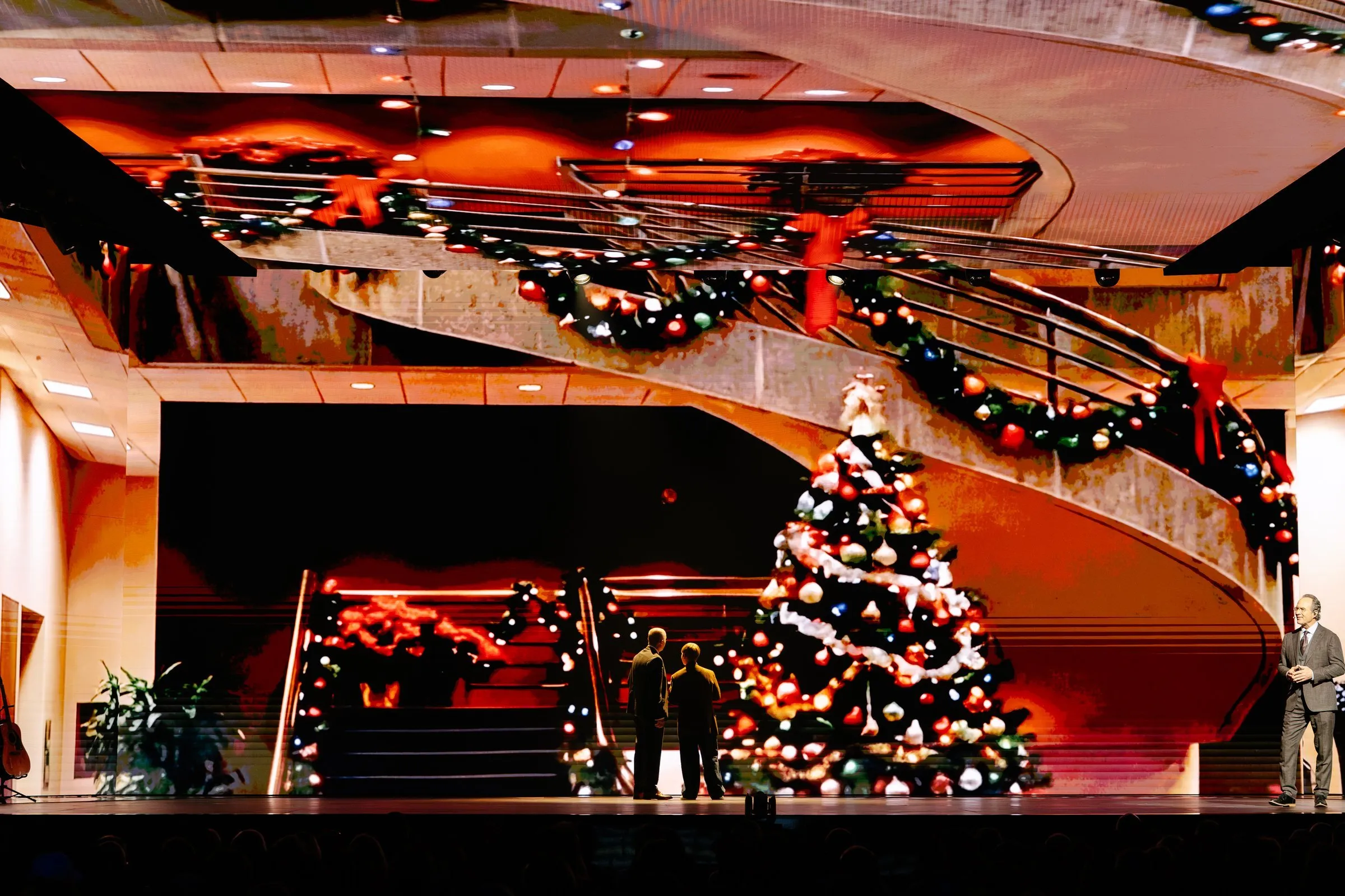 NEXT 2026 — holiday-themed stage scenic with wide curved LED screens above an interior staircase wrapped in garlands and a Christmas tree
