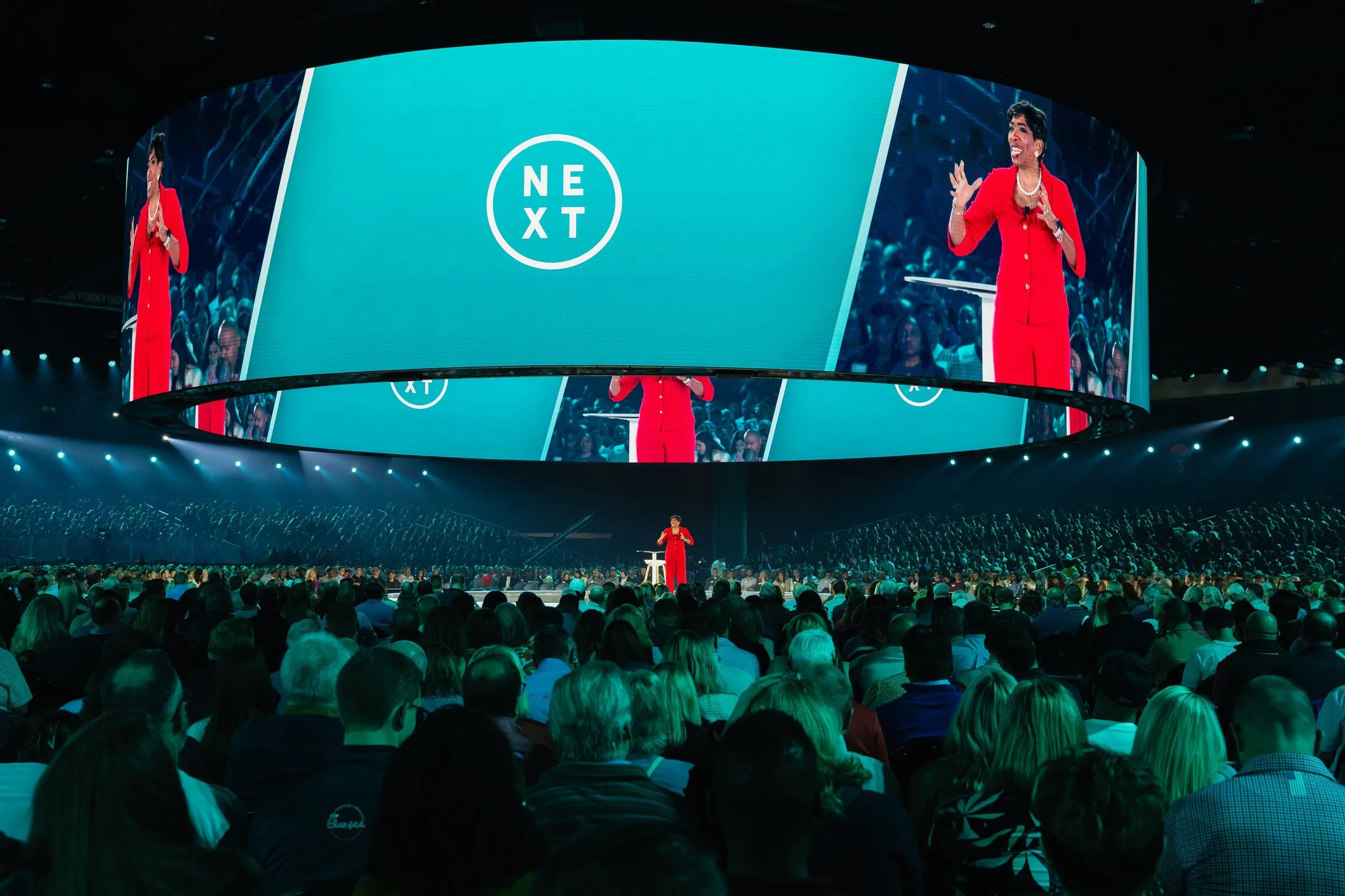 NEXT 2025 stage — circular NEXT mark on the suspended canopy with a speaker on a centered stage island
