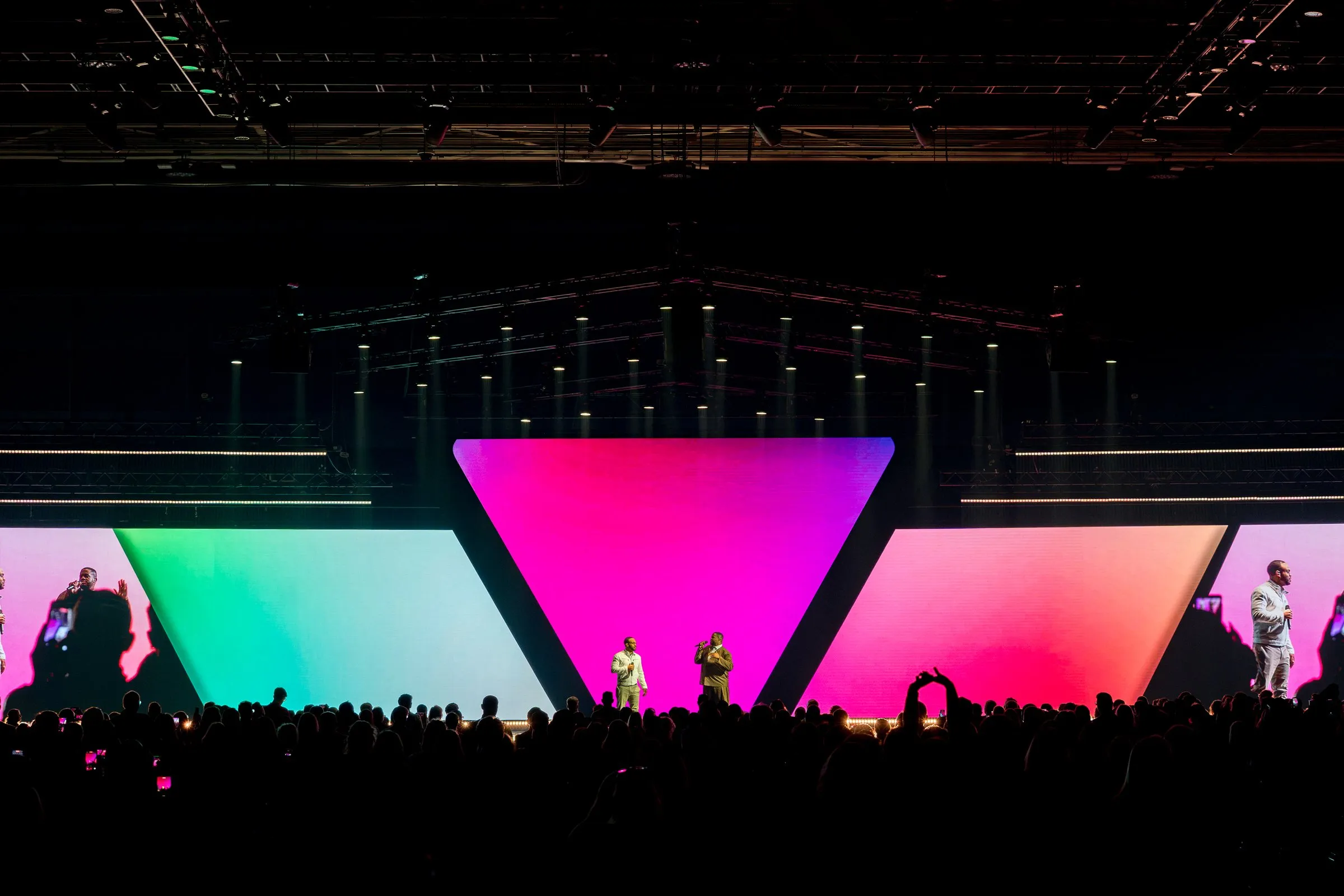 Vocalists on stage in front of a magenta-to-teal gradient look across the triangle center and angled wings