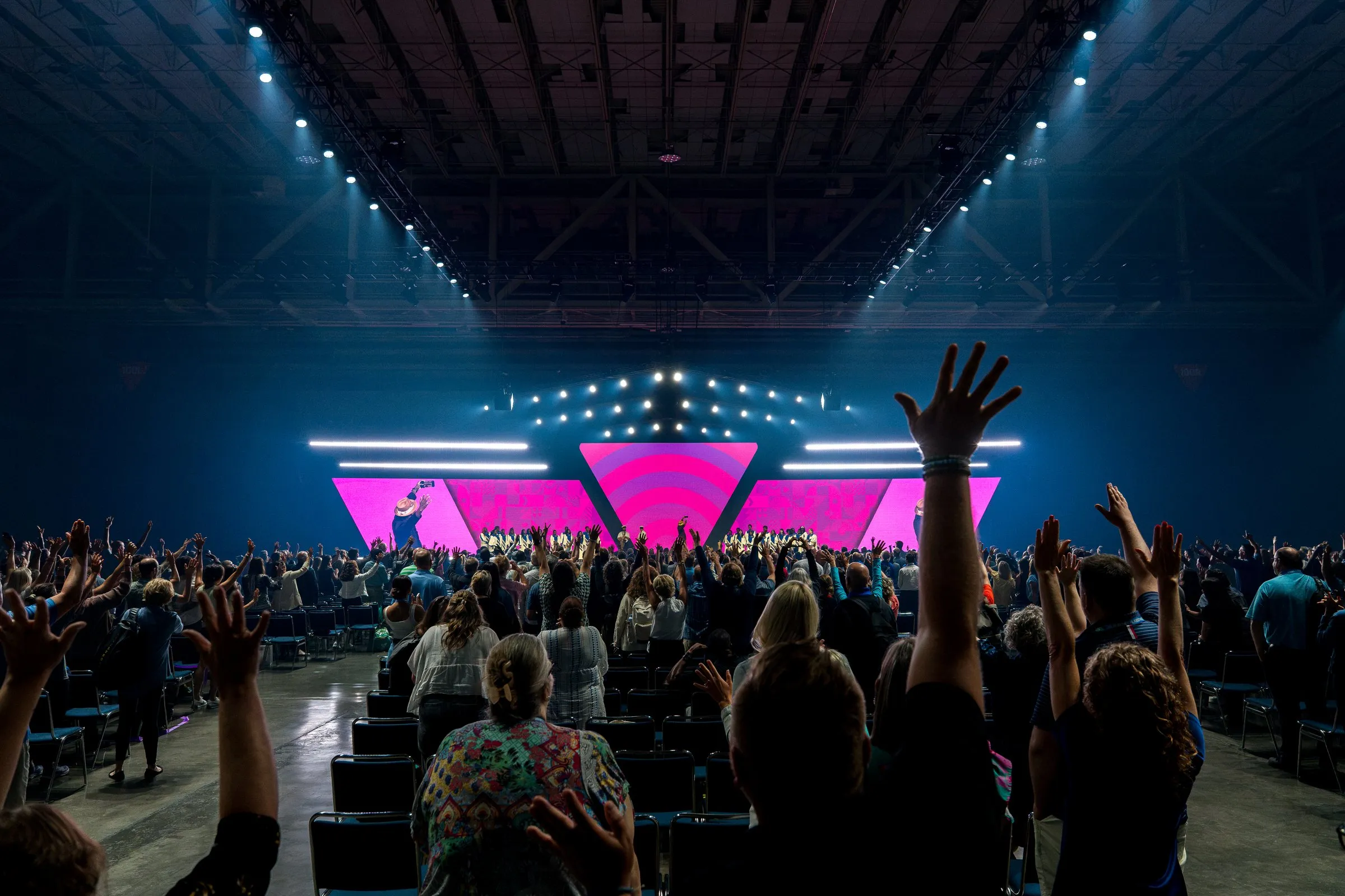The general session hall from the back — thousands of attendees with hands raised toward the pink LED build
