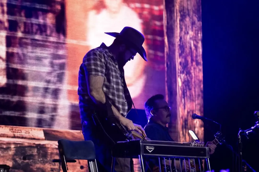 Sideman in a cowboy hat playing pedal steel with a distressed photo collage LED behind him