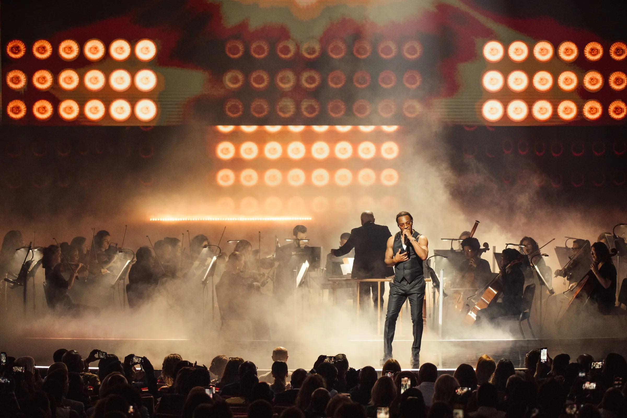 Beloved Benefit solo vocalist in front of a full classical orchestra with a Hollywood-style light-bulb wall illuminated behind