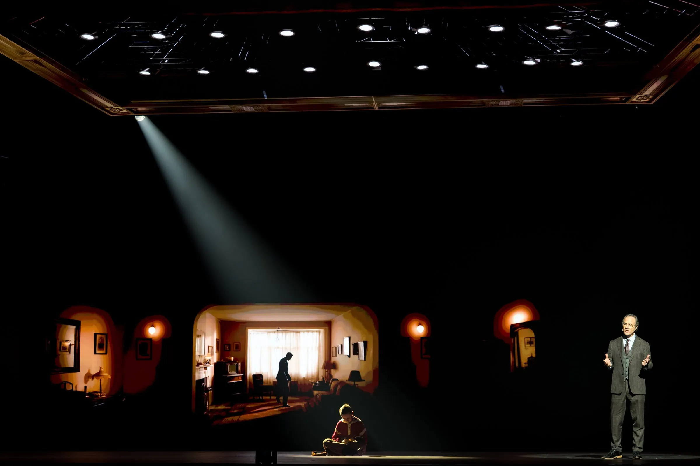 NEXT 2026 storytelling moment — single spotlight beam from a suspended grid frames a child sitting in a doorway, with a 1950s living room scene projected wide on the back LED wall