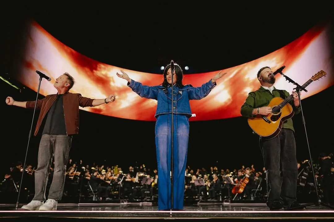 Beyond a Billion — three worship leaders performing on stage with a wide arched red-and-orange wave backdrop and orchestra below